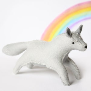 Può includere: Un giocattolo di unicorno in feltro grigio con un arcobaleno sullo sfondo. L'unicorno ha un corno e un occhio cucito.