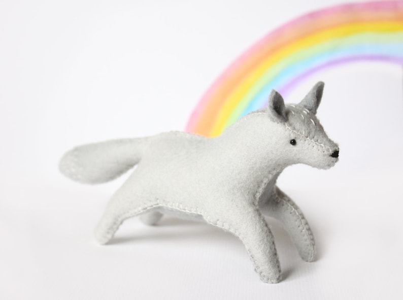May include: A gray felt unicorn toy with a rainbow in the background. The unicorn has a horn and a stitched-on eye.