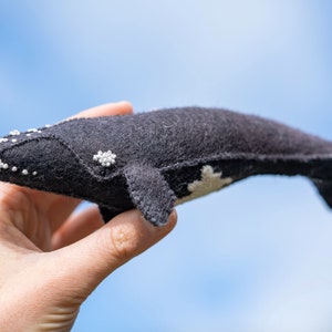 May include: A black felt whale with white bead accents, held in a hand against a blue sky.