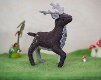 May include: A brown felt deer with gray antlers and white spots standing on a green felt surface. There are small red and white mushrooms in the background.