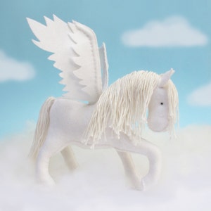 Peut inclure: Un cheval ailé en feutre blanc avec une longue crinière et une queue blanches. Le cheval est debout sur un fond de nuage blanc et moelleux.
