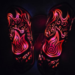 May include: Pair of black, illuminated leg warmers with a flame design. The warmers feature a cut-out pattern that glows with red and pink light, creating a fiery effect. The design includes swirling patterns and flame shapes, perfect for a costume or performance.