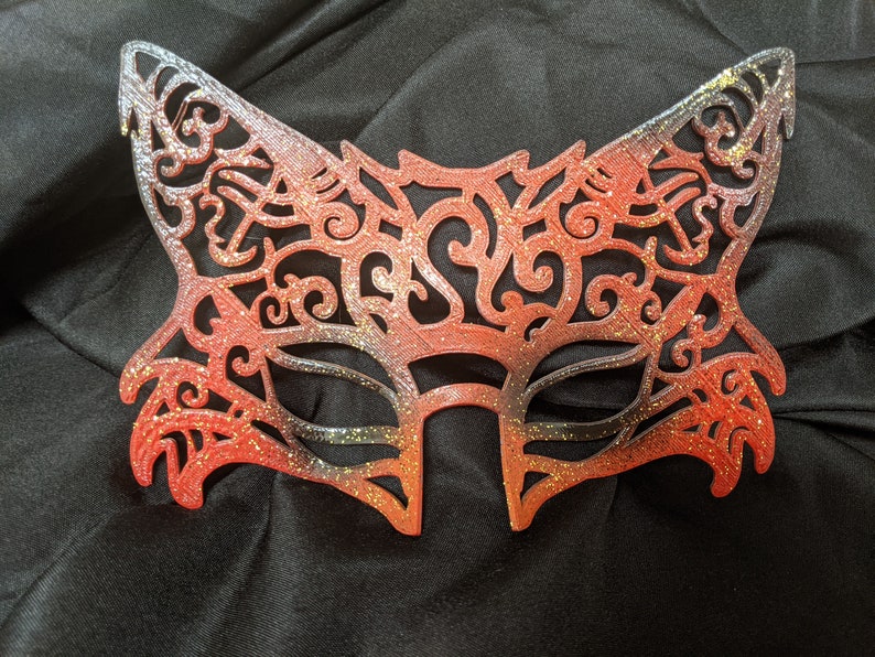 May include: A red and gold glitter cat mask with a delicate, intricate design. The mask is made of a lightweight material and has a comfortable fit.