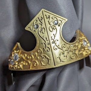 May include: A gold crown with a detailed floral design and clear rhinestones. The crown is made of a lightweight material and has a slightly curved shape.