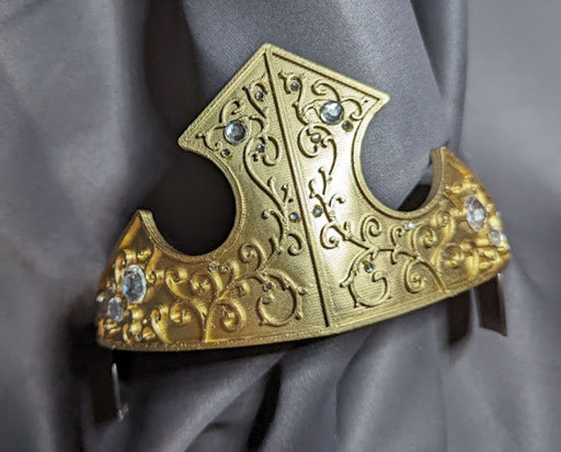 May include: A gold crown with a detailed floral design and clear rhinestones. The crown is made of a lightweight material and has a slightly curved shape.