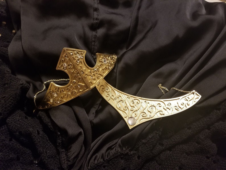 May include: A gold-toned crown with intricate floral designs and clear rhinestones. The crown is made of metal and has a delicate chain.