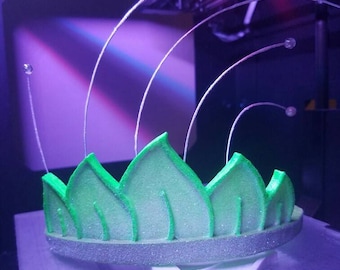 May include: A green and silver crown with a leaf design. The crown is being 3D printed and is suspended by thin wires.
