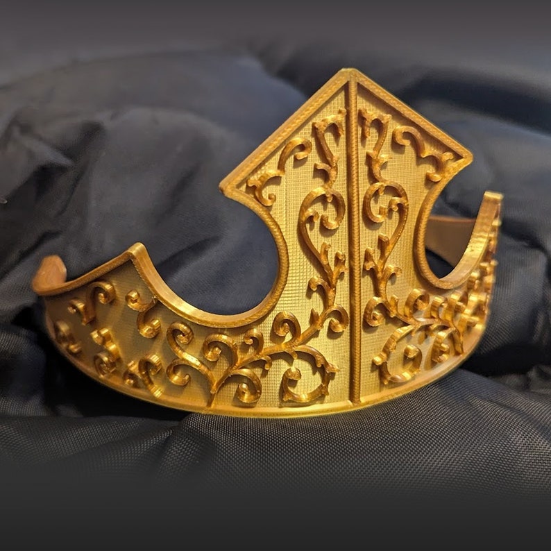 K&ouml;nnte beinhalten: Eine goldfarbene Krone mit einem detaillierten floralen Design. Die Krone ist aus einem glatten, gl&auml;nzenden Material gefertigt und hat eine spitze Spitze.
