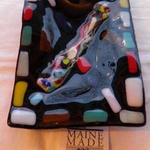 May include: A black glass dish with a colorful mosaic design. The dish is rectangular and has a raised edge. The mosaic design is made up of small pieces of glass in various colors, including red, blue, green, yellow, and white. The dish is labeled "Maine Made" and "America's Best".