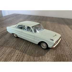 May include: A light blue die-cast model car with a classic design. The car features a detailed interior, chrome accents, and whitewall tyres. The model car is approximately 20 cm long.