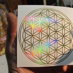 May include: A square white sticker featuring a gold-colored Flower of Life design. The geometric pattern has iridescent rainbow accents in the center. The sticker is held in a hand, with a blurred image in the background.