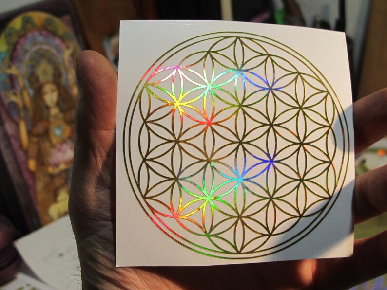 May include: A square white sticker featuring a gold-colored Flower of Life design. The geometric pattern has iridescent rainbow accents in the center. The sticker is held in a hand, with a blurred image in the background.