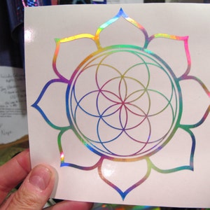 May include: A rainbow iridescent vinyl sticker featuring a flower with a geometric pattern inside. The flower has 12 petals and the geometric pattern is a circle with a flower-like design inside.