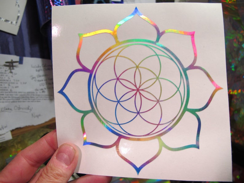 May include: A rainbow iridescent vinyl sticker featuring a flower with a geometric pattern inside. The flower has 12 petals and the geometric pattern is a circle with a flower-like design inside.