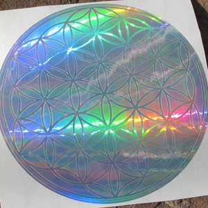 May include: A holographic sticker featuring the Flower of Life pattern. The circular design has a repeating geometric pattern with iridescent colors, reflecting light in a rainbow spectrum. The sticker is on a white background.