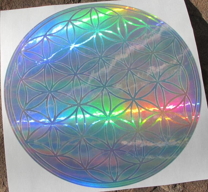 May include: A holographic sticker featuring the Flower of Life pattern. The circular design has a repeating geometric pattern with iridescent colors, reflecting light in a rainbow spectrum. The sticker is on a white background.