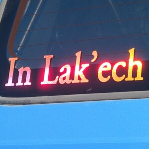 May include: A red, white, and orange sticker on a blue car window that reads "In Lak'ech".