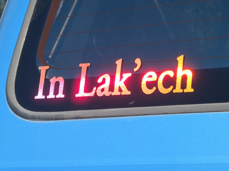 May include: A red, white, and orange sticker on a blue car window that reads "In Lak'ech".