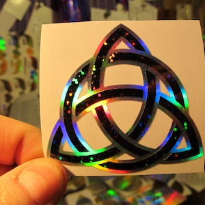 May include: A holographic sticker featuring a black and silver triquetra symbol with iridescent glitter. The triquetra is a Celtic knot symbol that represents the trinity.