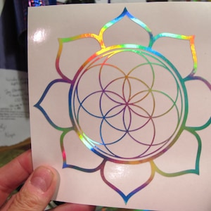 May include: A rainbow iridescent vinyl sticker featuring a flower with a geometric pattern inside. The flower has eight petals and the geometric pattern is a circle with a series of overlapping circles inside.
