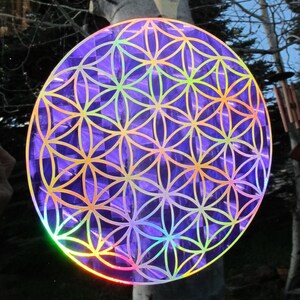 May include: A round, iridescent purple sticker featuring the Flower of Life pattern. The design has a geometric floral pattern with gold outlines. The sticker reflects a rainbow of colors in the light, creating a holographic effect.