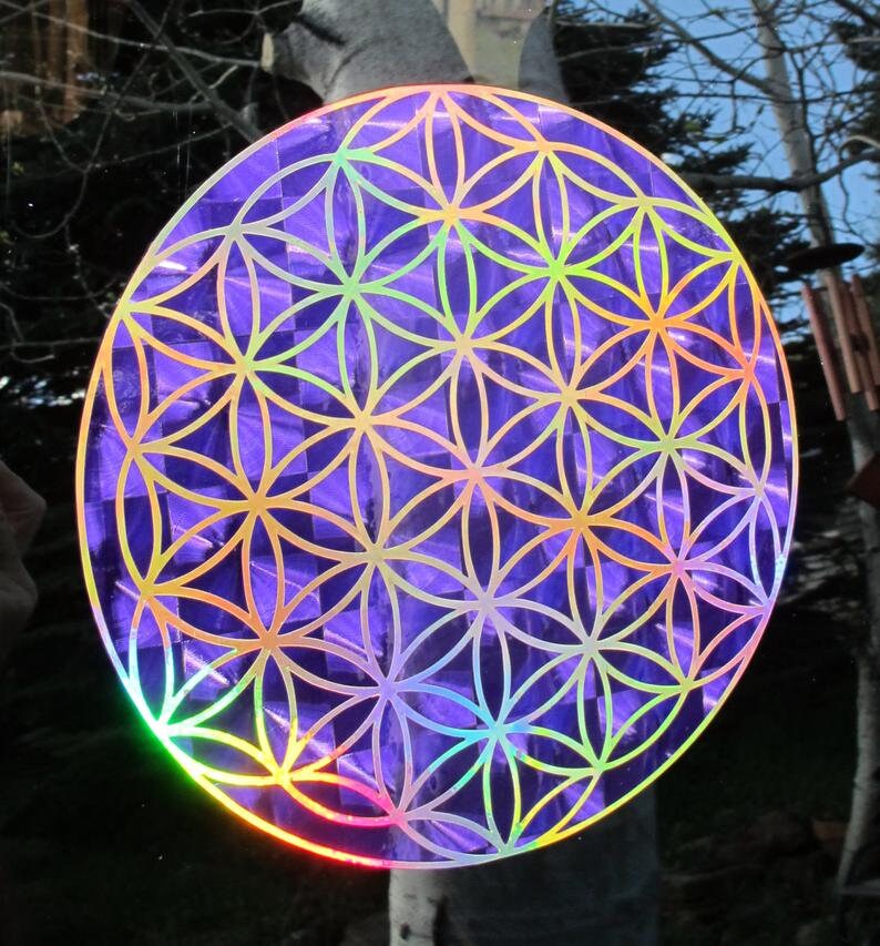 May include: A round, iridescent purple sticker featuring the Flower of Life pattern. The design has a geometric floral pattern with gold outlines. The sticker reflects a rainbow of colors in the light, creating a holographic effect.