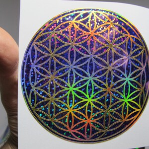 May include: A holographic sticker featuring the Flower of Life pattern. The design is a series of overlapping circles in a symmetrical pattern, with a shimmering, iridescent finish. The background is a dark blue with colorful glitter.