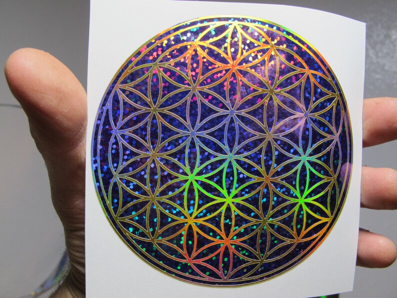 May include: A holographic sticker featuring the Flower of Life pattern. The design is a series of overlapping circles in a symmetrical pattern, with a shimmering, iridescent finish. The background is a dark blue with colorful glitter.