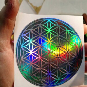 May include: A holographic sticker featuring a geometric flower of life design. The sticker has a reflective, iridescent finish with a spectrum of colors, including blue, green, and yellow. The sticker is on a white background.