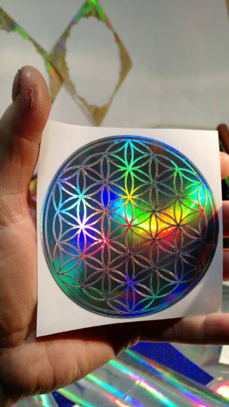 May include: A holographic sticker featuring a geometric flower of life design. The sticker has a reflective, iridescent finish with a spectrum of colors, including blue, green, and yellow. The sticker is on a white background.