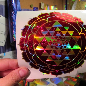 May include: A red and gold Sri Yantra sticker with a glittery finish. The sticker features a geometric pattern of triangles and a lotus flower design.
