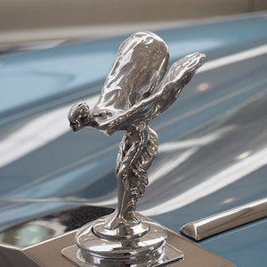 May include: A gleaming silver Spirit of Ecstasy hood ornament, a classic emblem of luxury cars. The sculpture shows a woman with flowing robes and outstretched arms. The Rolls-Royce logo is on the base.
