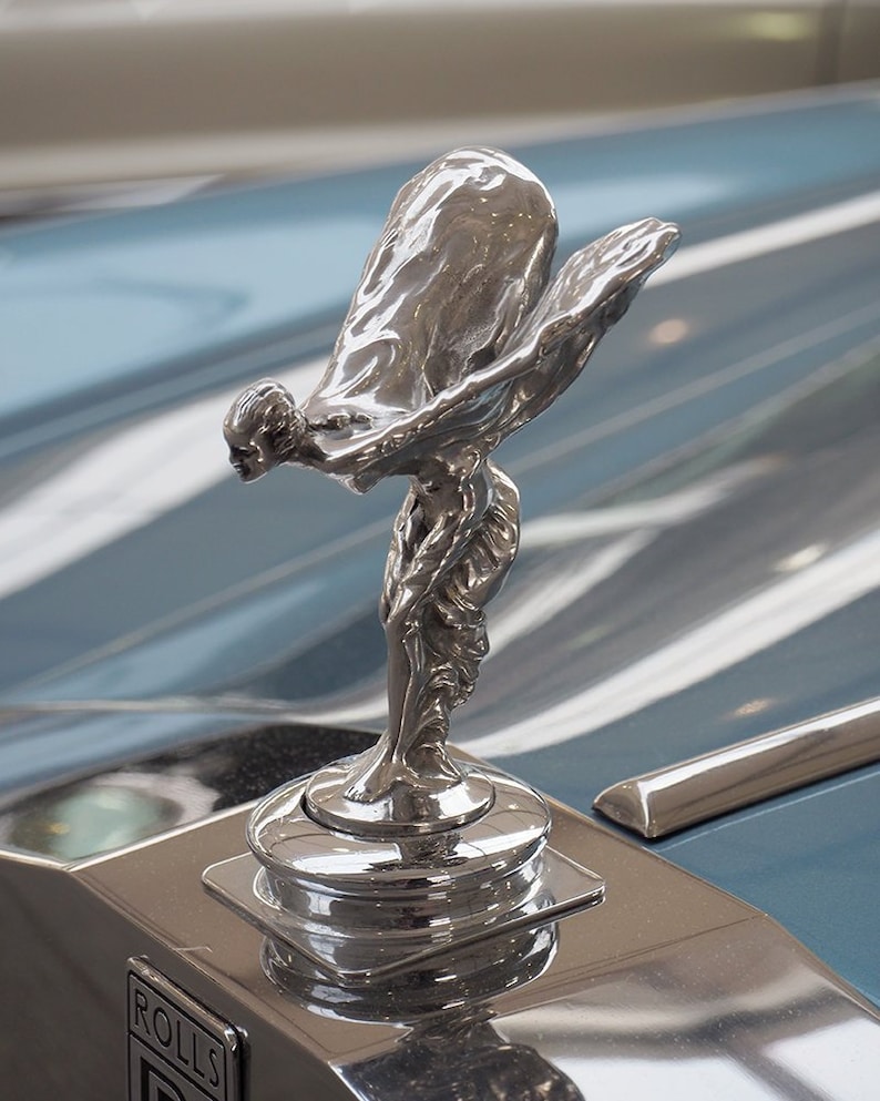 May include: A gleaming silver Spirit of Ecstasy hood ornament, a classic emblem of luxury cars. The sculpture shows a woman with flowing robes and outstretched arms. The Rolls-Royce logo is on the base.