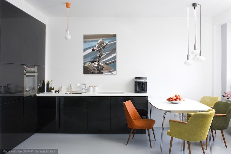 May include: A framed print of a silver Rolls-Royce hood ornament, the Spirit of Ecstasy, displayed in a modern kitchen. The kitchen features black cabinets, a white countertop, and a dining table with colourful chairs.