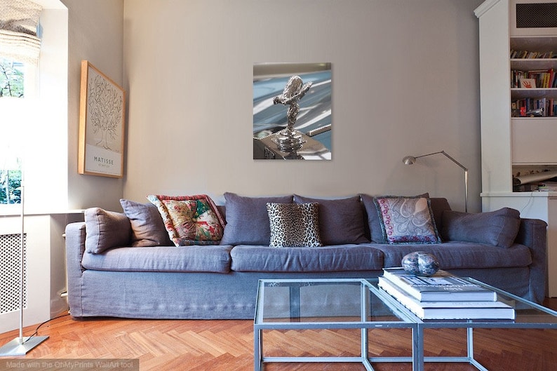 May include: A framed print of a silver car hood ornament, the Spirit of Ecstasy. The artwork is displayed on a wall above a grey sofa with decorative cushions. A glass coffee table and a framed tree print are in the foreground.