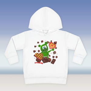 Puede incluir: Sudadera con capucha blanca con un oso de gominola verde que sostiene un corazón de chocolate. El oso está rodeado de corazones de chocolate y trozos de chocolate.