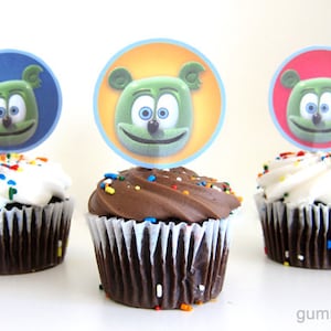 May include: Three chocolate cupcakes with white and chocolate frosting, topped with colourful sprinkles and gummy bear character cupcake toppers. The toppers are round and feature a green gummy bear with a blue, orange, and red background.