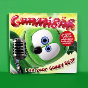 Könnte beinhalten: Ein grüner Gummibärchen-Cartoon-Charakter hält ein Mikrofon, mit dem Text "Gummibär" in Rot und "I am your Gummy Bear" in Weiß. Das Bild enthält auch einen roten Kreis mit dem Text "As seen on YouTube! Includes bonus tracks, videos, and extras!"