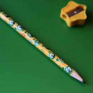 Puede incluir: Un lápiz amarillo con un diseño de personaje de dibujos animados verde y amarillo. El lápiz está sobre una superficie verde junto a un sacapuntas amarillo en forma de estrella.