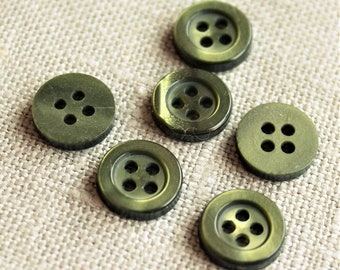 May include: Six olive green buttons with four holes each are arranged on a textured, light beige surface. The buttons have a slightly metallic sheen and a classic, round shape, suitable for various sewing projects.