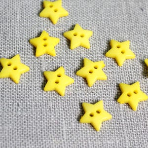 Könnte beinhalten: Eine Ansammlung leuchtend gelber sternförmiger Knöpfe mit jeweils zwei Löchern, verstreut auf einem strukturierten grauen Stoff. Diese kleinen, dekorativen Knöpfe eignen sich ideal für Näh- und Bastelprojekte.