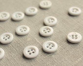May include: A collection of white plastic buttons with four holes each. The buttons are arranged in a scattered pattern on a light beige fabric surface.
