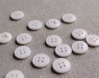 May include: A collection of white plastic buttons with four holes each. The buttons are arranged in a random pattern on a light gray fabric surface.