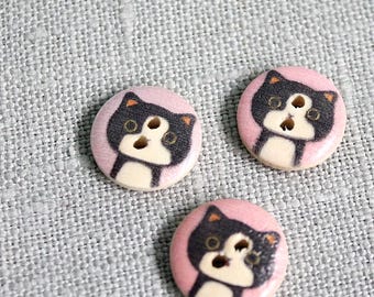 Puede incluir: Tres botones redondos de madera con un diseño de cara de gato. Cada botón presenta un fondo rosa con una ilustración de gato en blanco y negro. Los botones miden aproximadamente 2,5 cm de diámetro y tienen dos agujeros para coser.