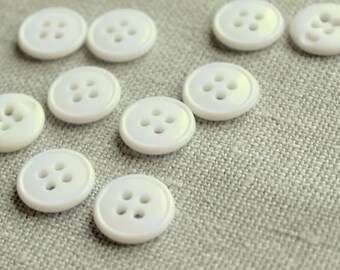 May include: Ten white plastic buttons with four holes each. The buttons are arranged in a scattered pattern on a light gray fabric background.