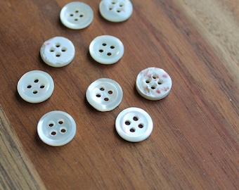 May include: A collection of round, off-white buttons with four holes each, scattered on a wooden surface. Some buttons have a pearlescent sheen, while others show slight variations in texture and color. The buttons are a classic sewing or craft supply.
