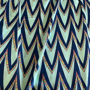 May include: A fabric with a repeating chevron pattern. The design features navy blue chevrons on a light green background, with colorful striped accents in yellow, pink, and red. The fabric has a slight sheen.