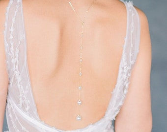 May include: A long, delicate gold chain with pearls hangs from the back of a person's neck. The chain is adorned with small pearls and larger pearls at the bottom.