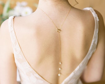 May include: A bride wearing a white lace wedding dress with a low back. She is wearing a delicate gold necklace with pearls hanging from it.
