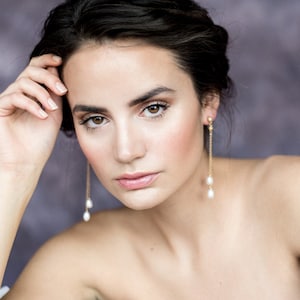Puede incluir: Una mujer con cabello oscuro lleva un vestido blanco sin tirantes y largos pendientes de perlas. Ella está mirando a la cámara con una expresión suave.